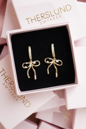 Therslund Smykker Bow ørestikker Bow ørestikkerne er fremstillet i forgyldt messing , et par elegante guldfarvet øreringe formet som sløjfer, meget feminint og let Øverst sidder lodret stav med små sten under staven hænger selve sløjfen i en blød og organisk formsprog med  fine, rinde buer og båndender der falder let ned, romantisk og sødt. Disse ørestikker er en ideel gaveidé eller en stilfuld detalje, der kan løfte ethvert outfit og tilføje et strejf af elegance. Modernet og minimalistisk, brug den til et bryllup, date night eller når du føler der skal lidt extra ind i hverdagen. Alle vores smykker er nikkel- og blyfri og lever op til dansk lovgivning. Smykkerne er fremstillet i messing eller rustfrit stål og kombineres ofte med glasperler eller halvædelsten for at skabe unikke og holdbare designs.