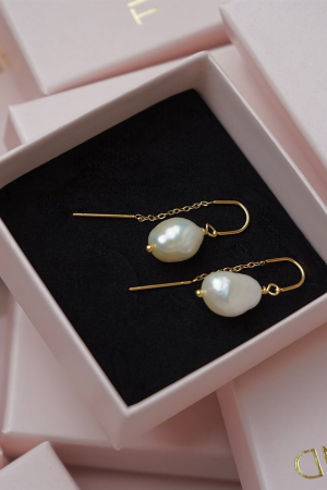 Therslund Smykker – Celina ØreringeCelina-øreringene er fremstillet i forgyldt messing og kombinerer elegance med et minimalistisk design. De er udsmykket med barokke ferskvandsperler, der med deres let ujævne, organiske form skaber et unikt og naturligt udtryk. Øreringene har guldfarvede kroge med en fin kædedetalje, som tilføjer lethed og et moderne, svævende look. Perlernes bløde glans står i smuk kontrast til det varme guld og giver øreringene et tidløst, feminint præg. Disse øreringe er alsidige og passer perfekt til: • Festlige lejligheder som bryllupper eller middage • Et sofistikeret hverdagslook • En enkel kjole eller en klassisk skjorte   Designet forener klassisk perleelegance med et moderne, let asymmetrisk udtryk, hvilket gør dem til en stilfuld detalje, der kan løfte ethvert outfit. De er den ideelle gaveidé eller et elegant tilbehør til både hverdag og særlige anledninger. Alle vores smykker er nikkel- og blyfri og overholder dansk lovgivning. Smykkerne fremstilles i messing eller rustfrit stål og kombineres ofte med glasperler eller halvædelsten for at skabe unikke og holdbare designs.