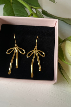 Therslund Smykker Elina Øreringe Øreringene har guldfarvede, som tilføjer lethed og et moderne, svævende look. designet er en smuk sløjfe der tilfører en kontrast til det varme guld og giver øreringene et tidløst, feminint præg. Disse øreringe er alsidige og passer perfekt til: Festlige lejligheder som bryllupper eller middage Et sofistikeret hverdagslook En enkel kjole eller en klassisk skjorte Designet forener klassisk og let asymmetrisk udtryk, hvilket gør dem til en stilfuld detalje, der kan løfte ethvert outfit. De er den ideelle gaveidé eller et elegant tilbehør til både hverdag og særlige anledninger. Alle vores smykker er nikkel- og blyfri og overholder dansk lovgivning. Smykkerne fremstilles i messing eller rustfrit stål og kombineres ofte med glasperler eller halvædelsten for at skabe unikke og holdbare designs.